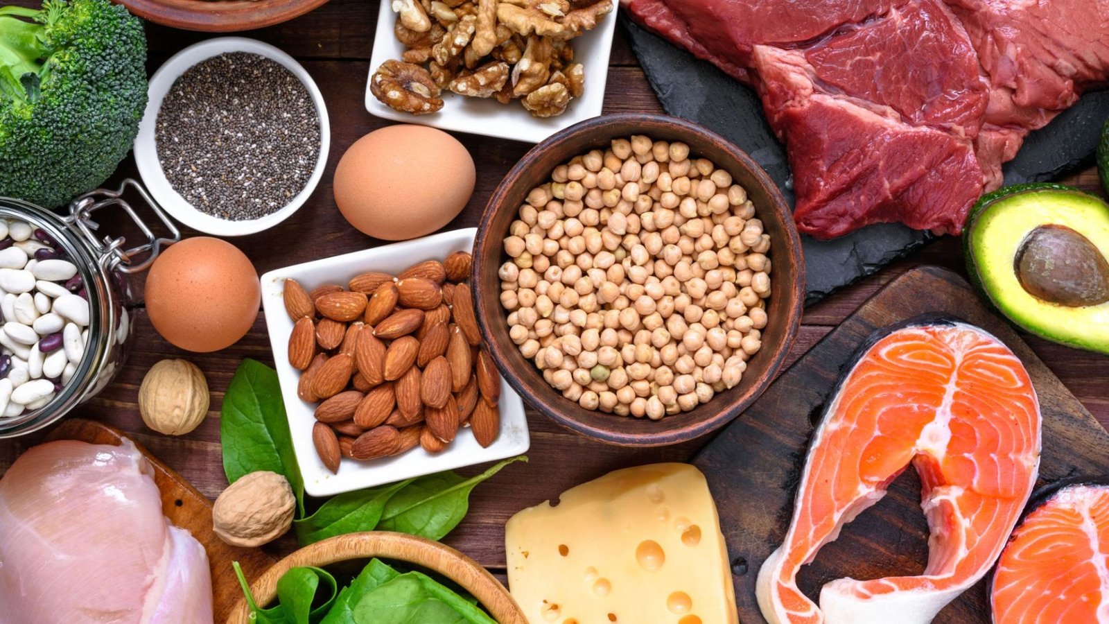 Assorted protein and produce on a wooden table: beef, salmon, eggs, cheese, nuts, seeds, legumes, avocado, broccoli and greens.
