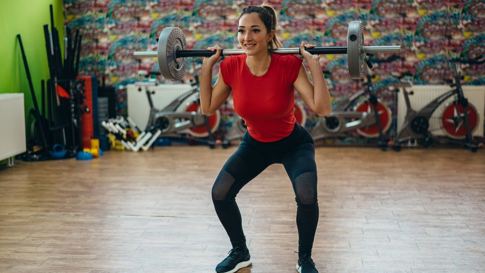 Woman performing a barbell back squat in a gym, wearing a red shirt and black leggings.