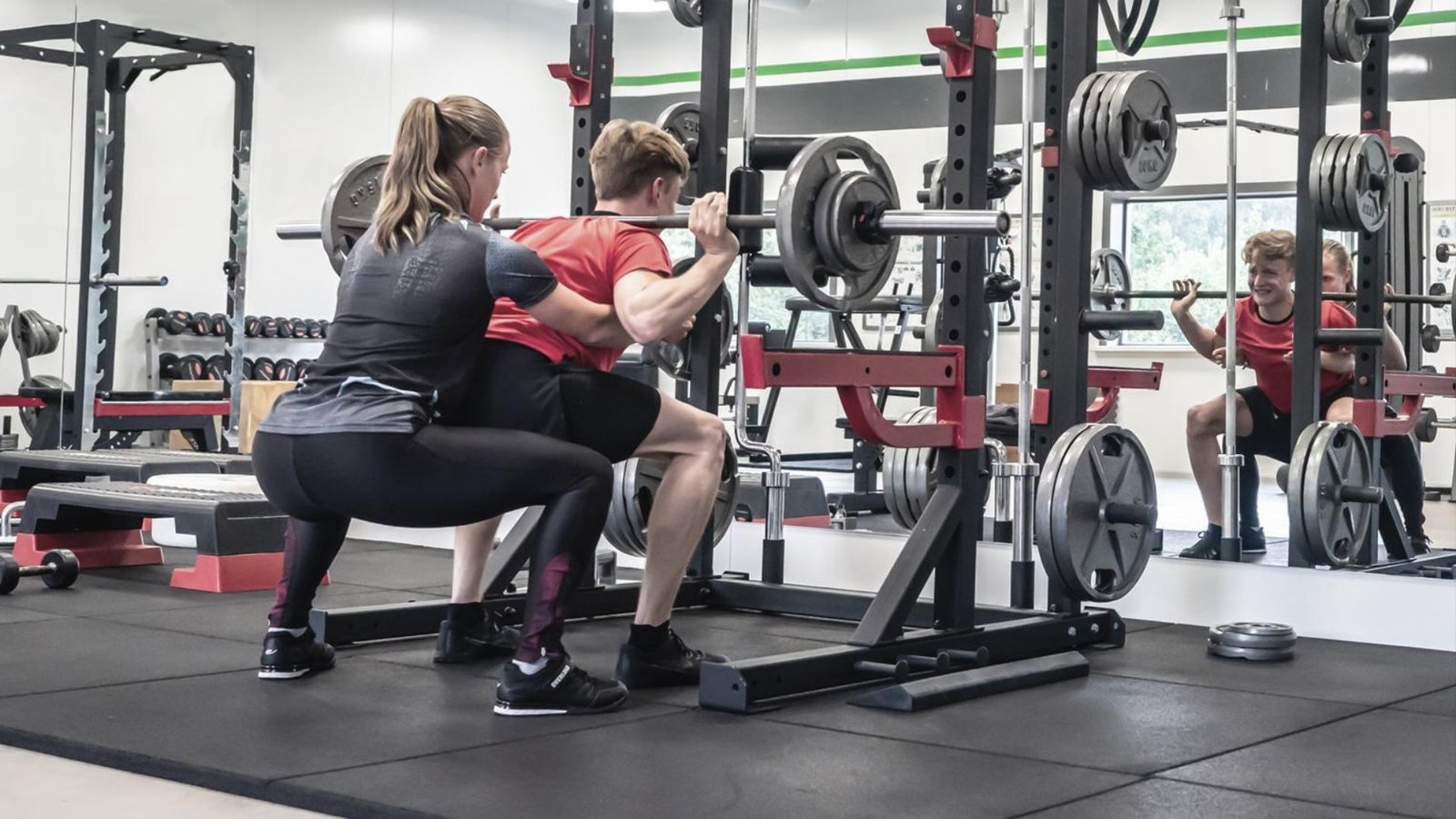 Man squats with a loaded barbell while a woman spots him in a gym squat rack with weight plates nearby in view.